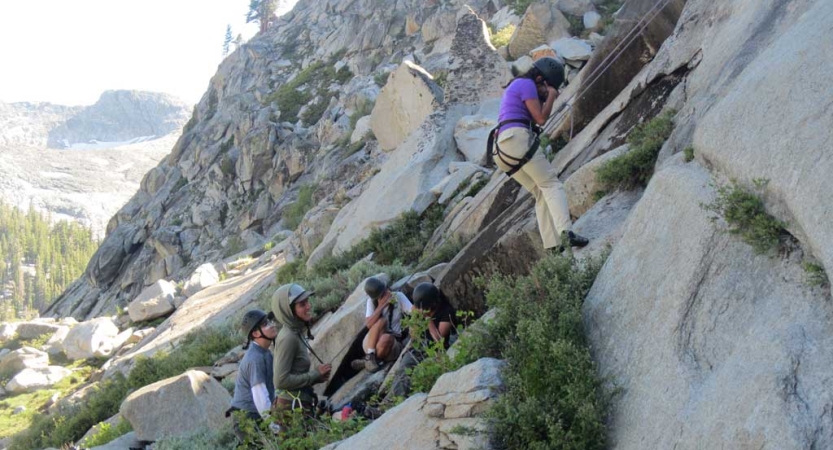 A group of students stand on the ground below a rock climber who is wearing safety gear and is secured by ropes. The people on the ground seem to be encouraging the climber.